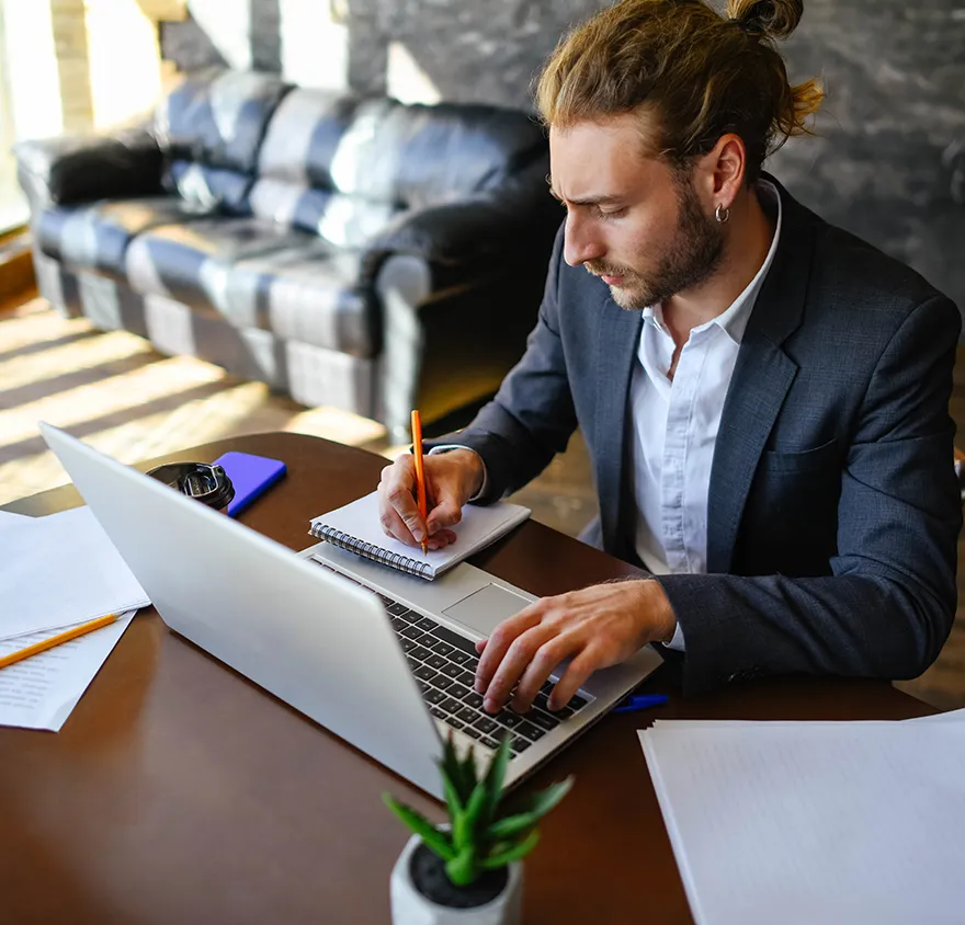 Man in his 30's in a suit writing a note at his desk